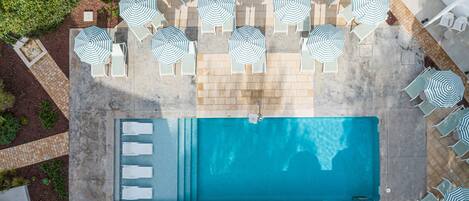 An aerial view of a swimming pool surrounded by lounge chairs and umbrellas, with a tiled deck and greenery.