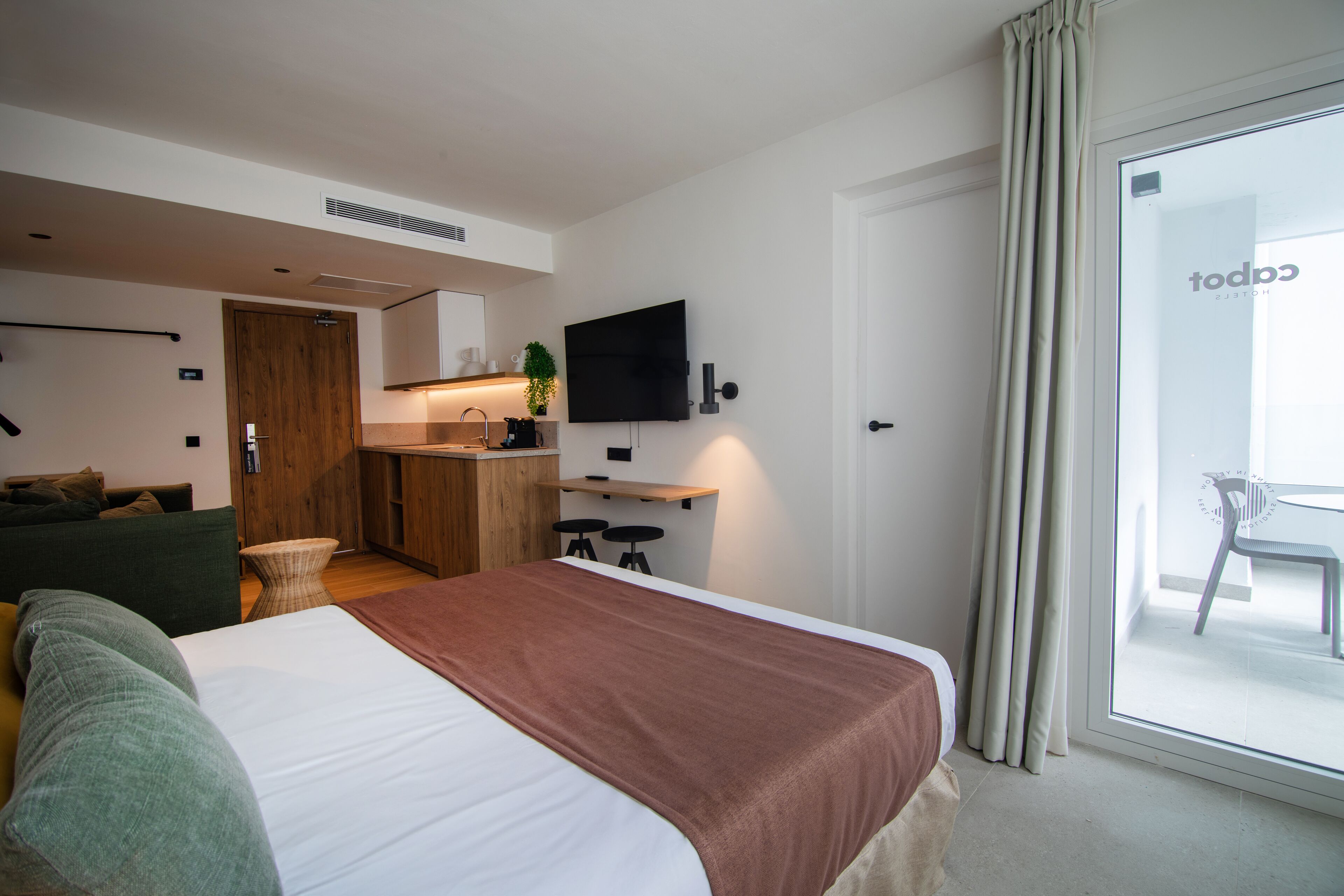 Una habitación de hotel moderna con una cama grande, una pequeña kitchenette, un televisor de pantalla plana y un puerta corredera de vidrio al balcón.