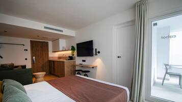 Una habitación de hotel moderna con una cama grande, una pequeña kitchenette, un televisor de pantalla plana y un puerta corredera de vidrio al balcón.