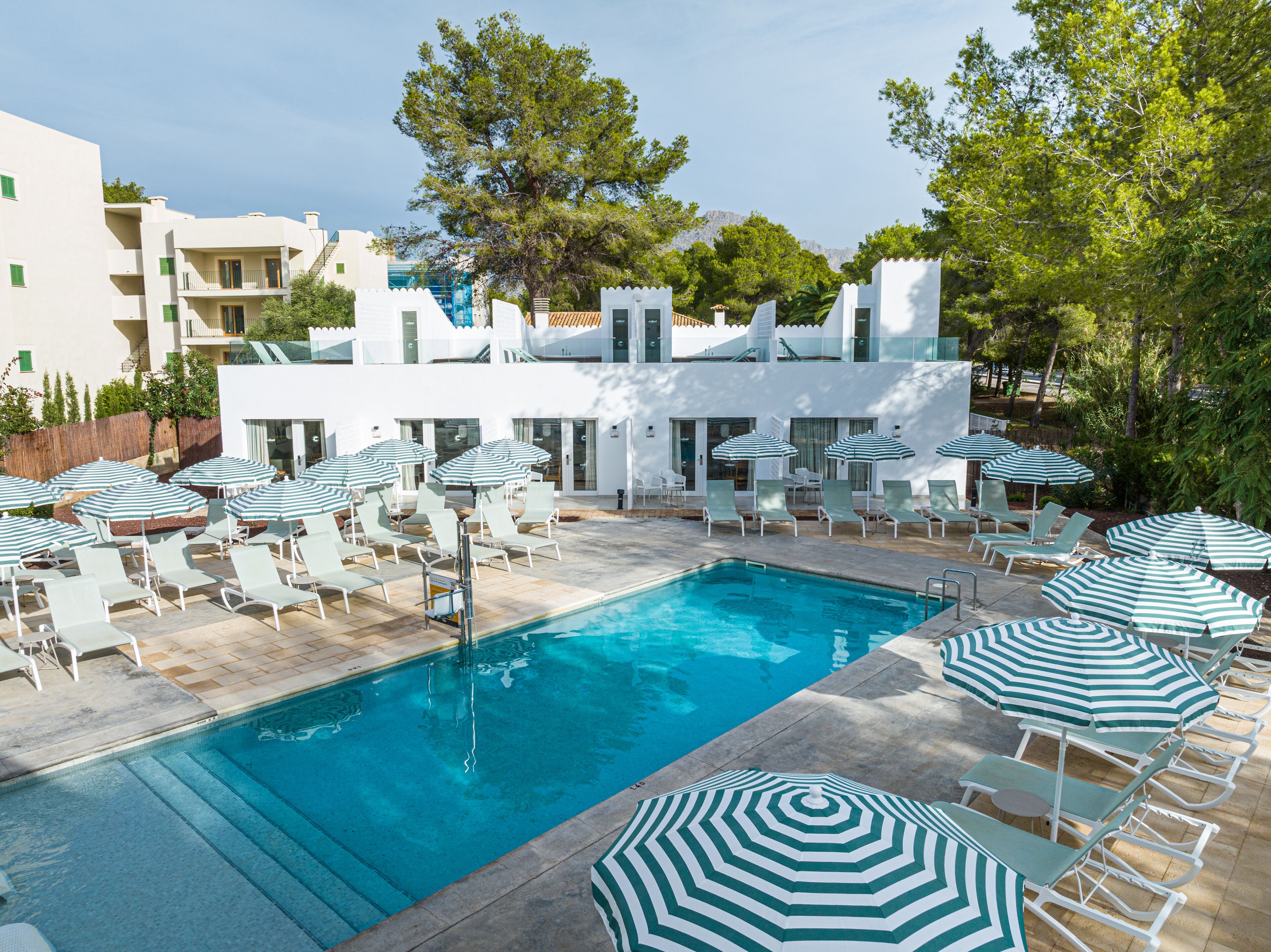 Un espace piscine doté de chaises longues et de parasols à rayures, entouré de bâtiments blancs et d’arbres.