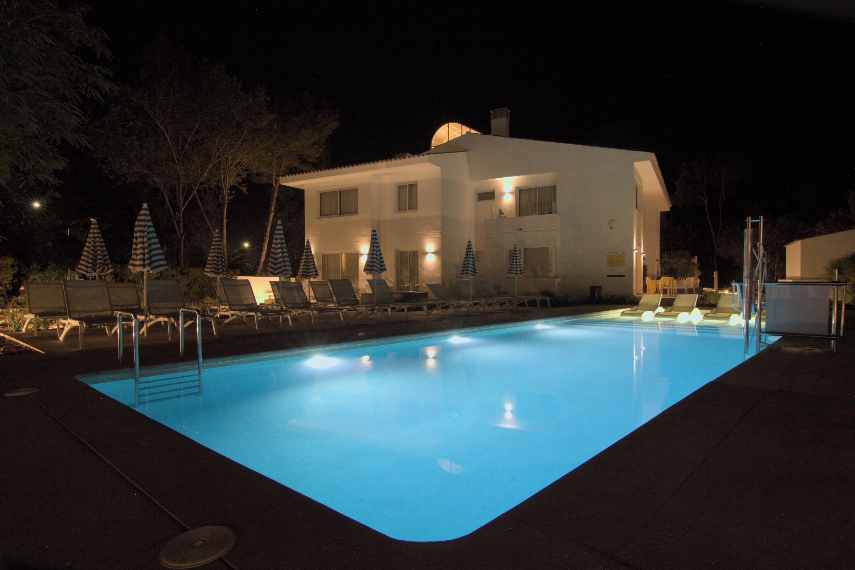 Une piscine entourée de chaises longues et de parasols, elle-même bordée d’un bâtiment doté de nombreuses fenêtres, le tout dans une ambiance nocturne.