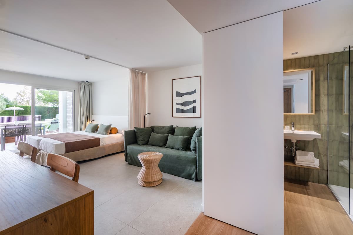 A modern hotel room with a large bed, a sofa, a dining table, and a glass shower area.