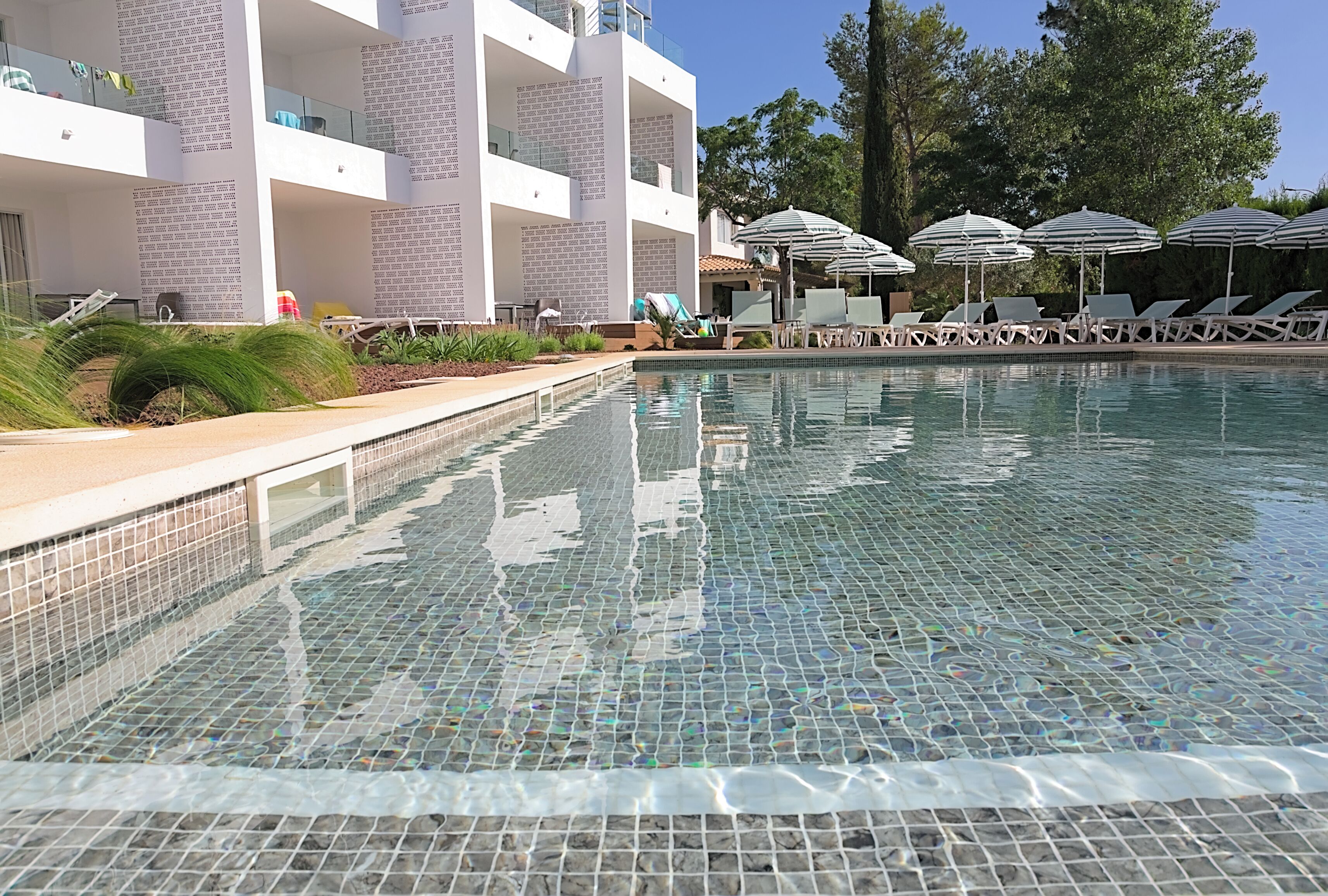 Una piscina con diseño de mosaicos, rodeada de sillas de descanso y sombrillas, y un edificio de varios pisos con balcones al fondo.