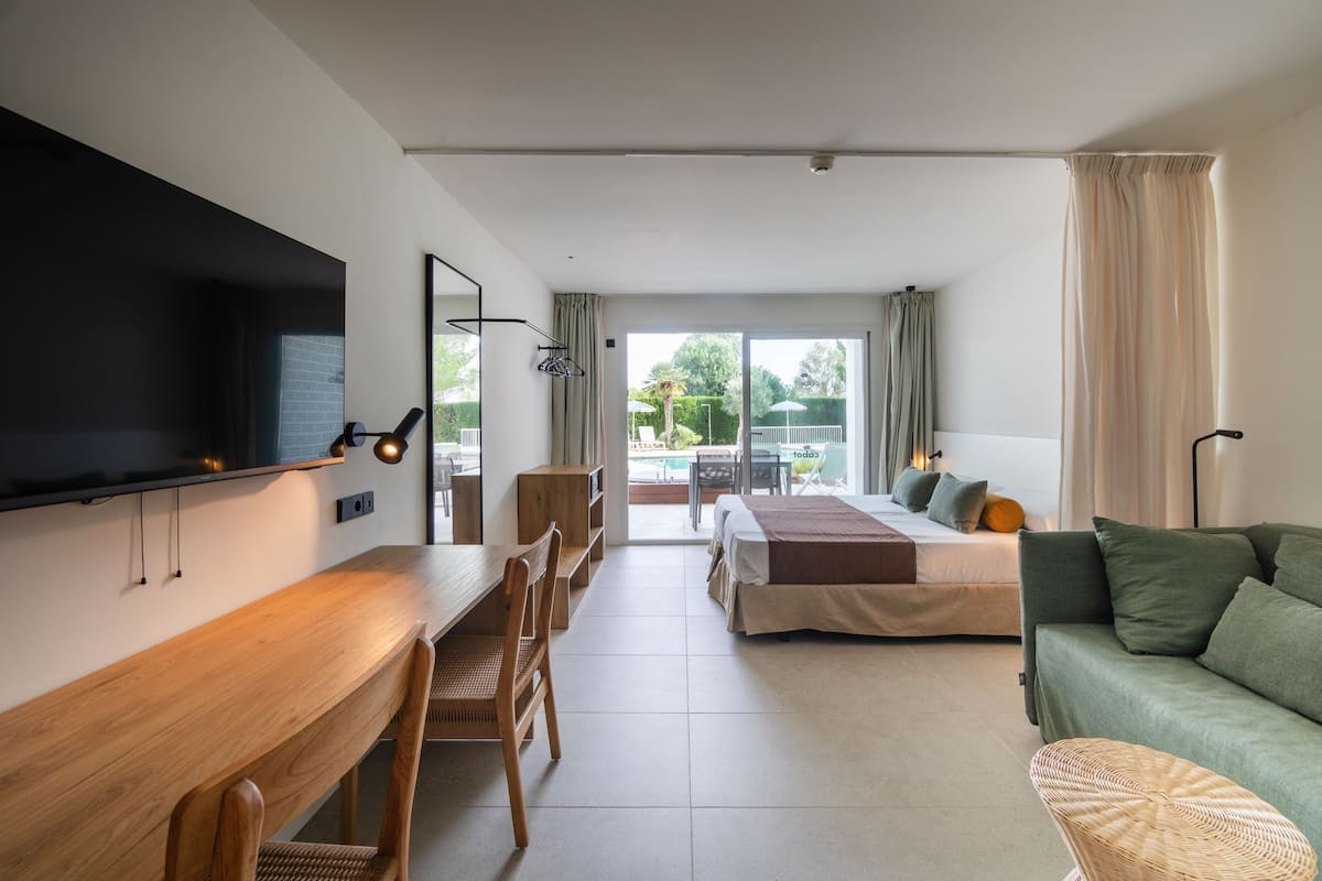 A modern hotel room with a wooden desk, a bed with green bedding, a green sofa, and a view of a pool and garden.