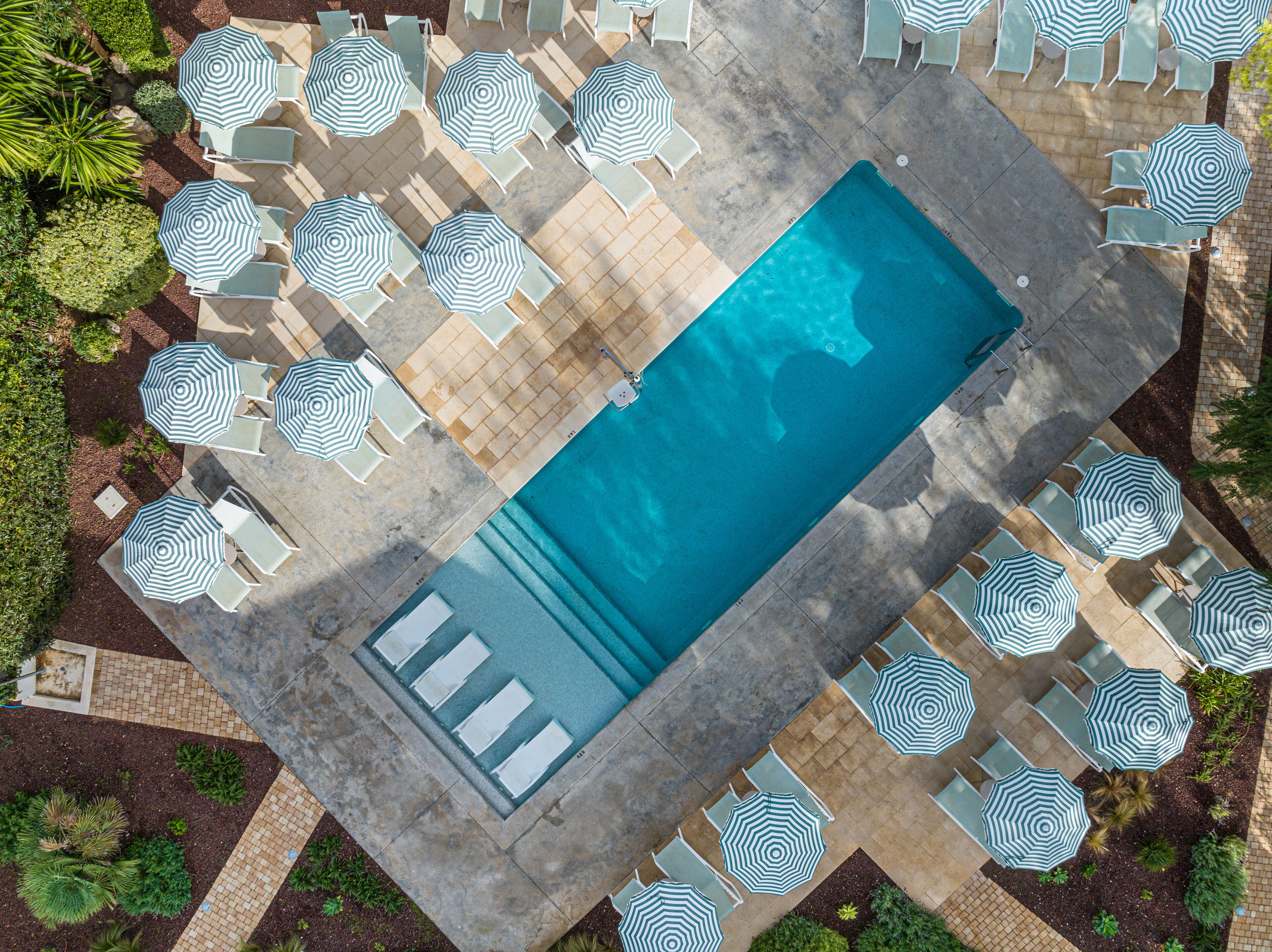 Ein Luftbild eines rechteckigen Schwimmbeckens, umgeben von Liegestühlen und gestreiften Sonnenschirmen, mit einer gefliesten Liegefläche.