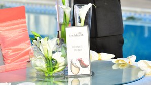 A waiter holding a tray with glasses and a menu at a poolside table with floral arrangements and a sign reading "The Manor Drinks Menu".