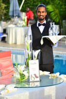 A waiter holding a tray with glasses and a menu at a poolside table with floral arrangements and a sign reading "The Manor Drinks Menu".