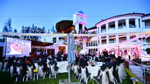An outdoor event with tables and chairs arranged for guests, a large screen displaying an image, and a building with balconies in the background.