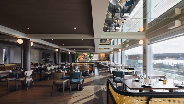 A spacious restaurant with multiple dining areas, large windows offering views of the city, and a modern interior design. - Hôtel Versō (Magog)