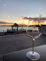 Una copa de vino sobre una mesa, con vista a un atardecer sobre el agua y un pabellón al fondo.