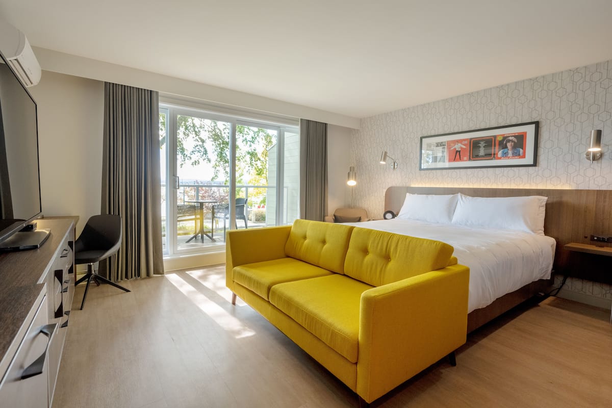 A hotel room with a bed, a yellow sofa, a desk with a chair, and a large window with a view of the outdoors.