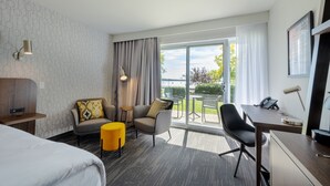 A hotel room with a bed, a desk with a telephone, a chair, a small table, and a view of the outdoors. - Hôtel Versō (Magog)