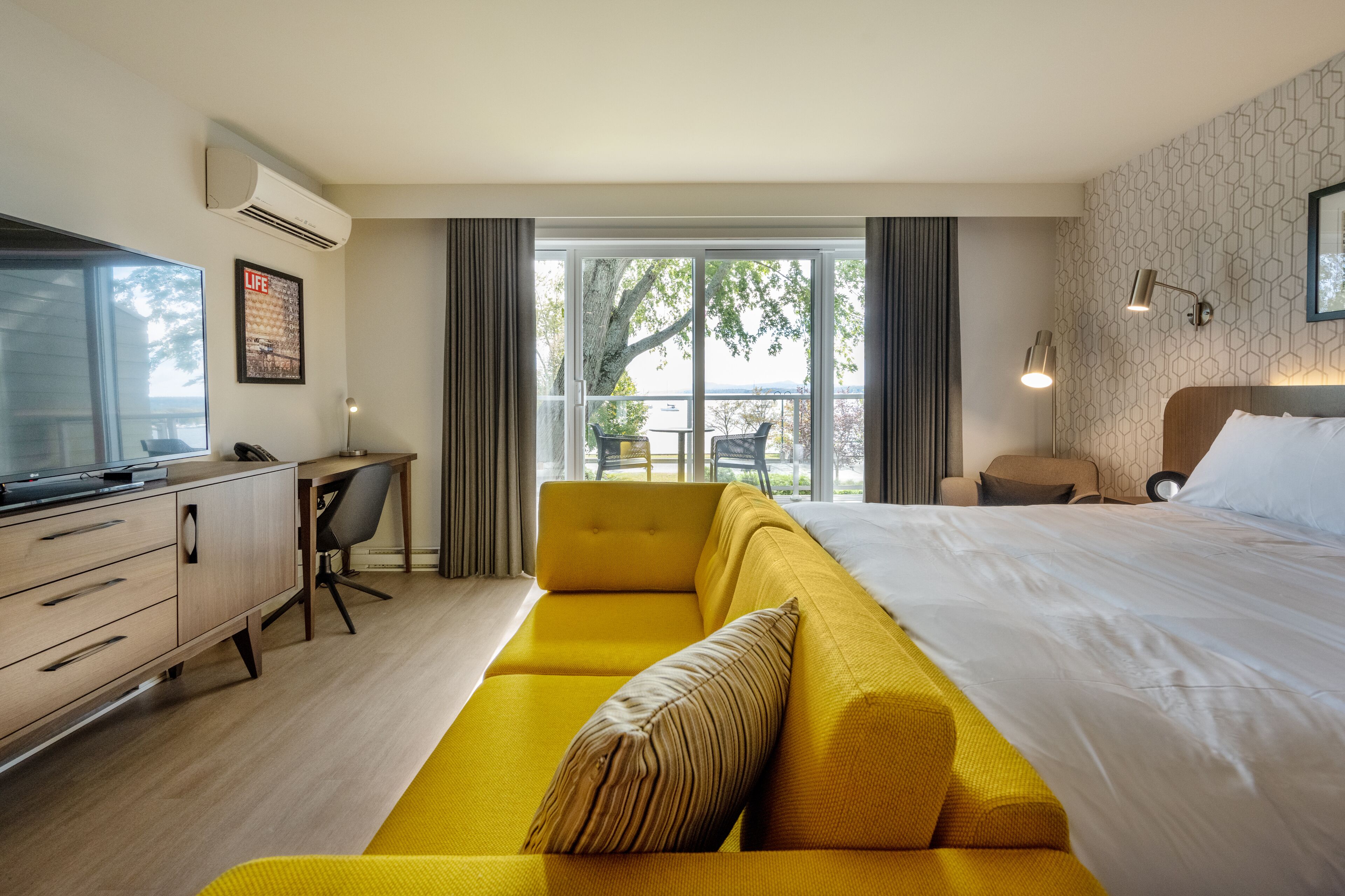 A modern hotel room with a large bed, a yellow sofa, a desk with a computer, and a view of the outdoors.