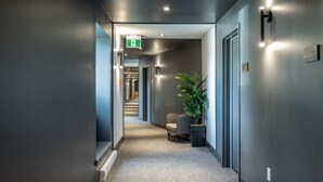 A modern hallway with a carpeted floor, a potted plant, a chair, and a door with an emergency exit sign. - Hôtel Versō (Magog)