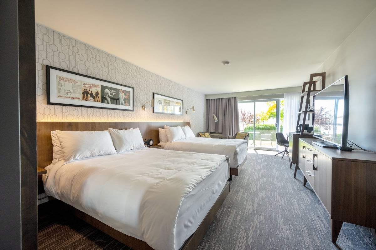 A hotel room with two beds, a desk, a chair, and a large window with a view of trees.