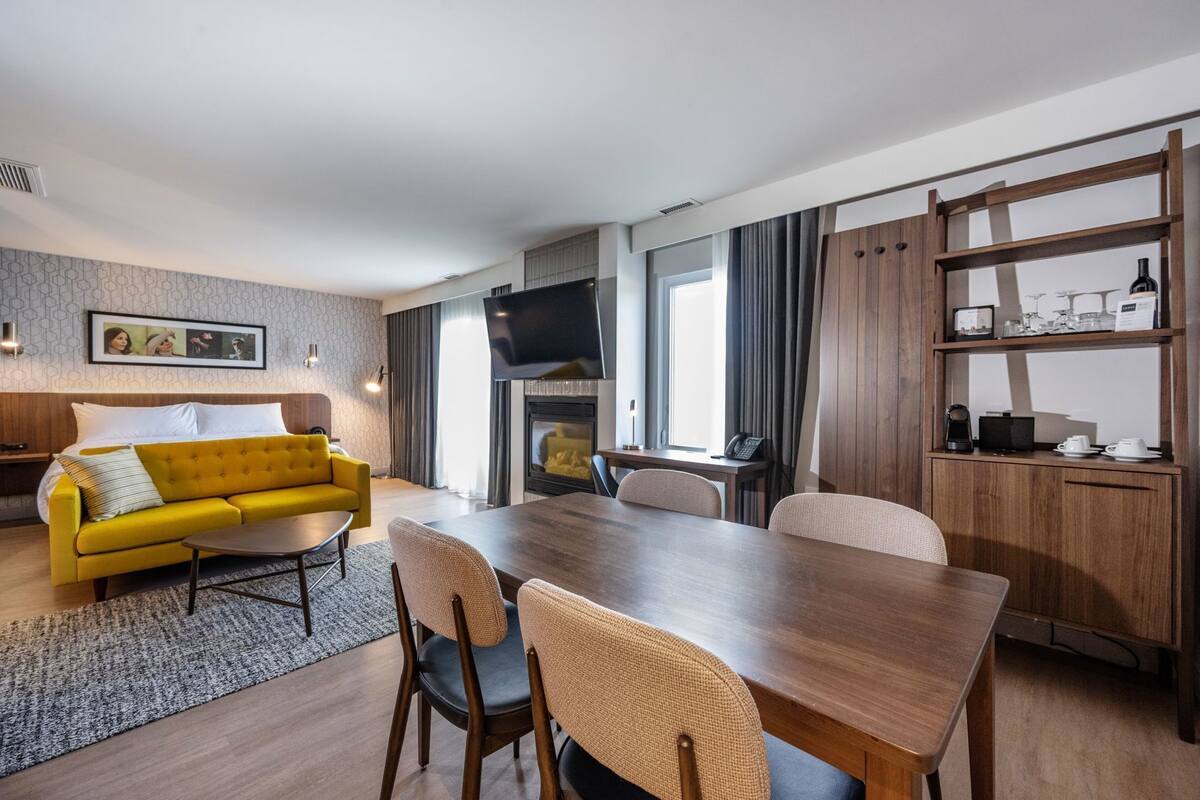 A modern hotel room with a yellow sofa, a wooden dining table, and a fireplace.