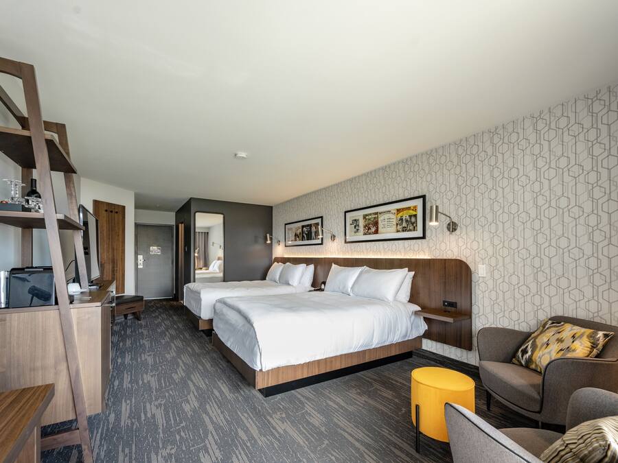 A hotel room with two beds, a desk, a chair, a small table, and a yellow ottoman.