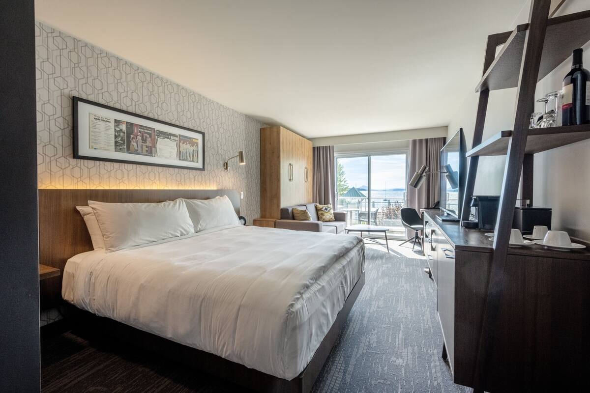 A modern hotel room with a large bed, a desk, a chair, and a balcony with a view.