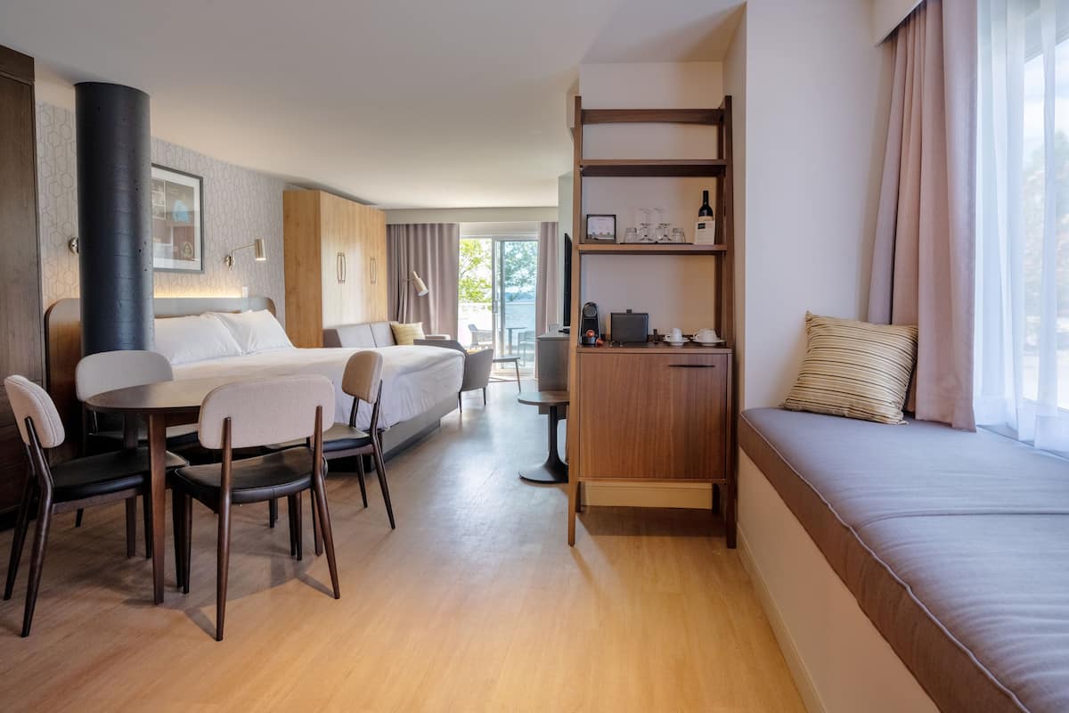 A modern hotel room with a bed, a sofa, a dining table with chairs, a TV, and a coffee machine.