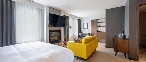 A modern hotel room with a large bed, a yellow sofa, a TV, a fireplace, and a kitchenette.