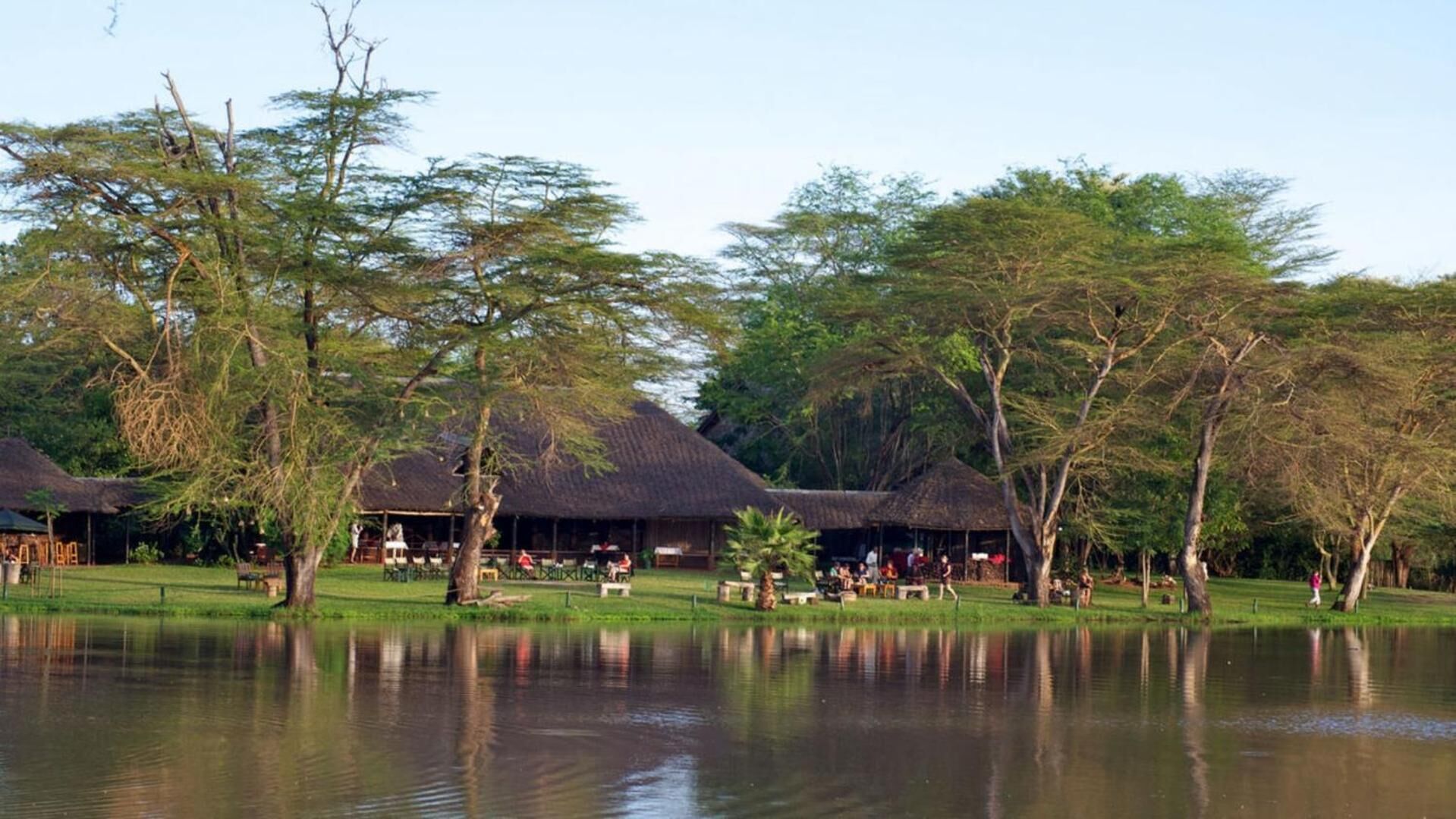A lakeside resort with thatched-roof huts, open grassy areas, and trees.