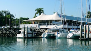A marina with several boats docked, a covered area with a white canopy, and a rocky shoreline in the background. - The Beach Place (Yorkeys Knob)