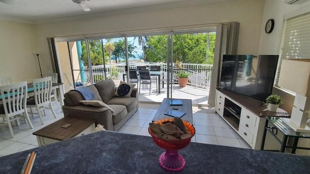 A living room with a sofa, coffee table, TV, and a balcony with outdoor furniture. - The Beach Place (Yorkeys Knob)