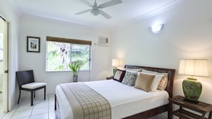 A neatly arranged bedroom with a bed, a chair, a nightstand, a window with a view of greenery, and a ceiling fan. - The Beach Place (Yorkeys Knob)