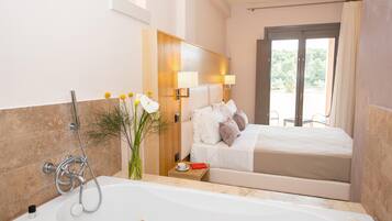 A modern hotel room with a large bathtub, a bed with white linens, a wooden headboard, and a view of the outdoors through a sliding door.