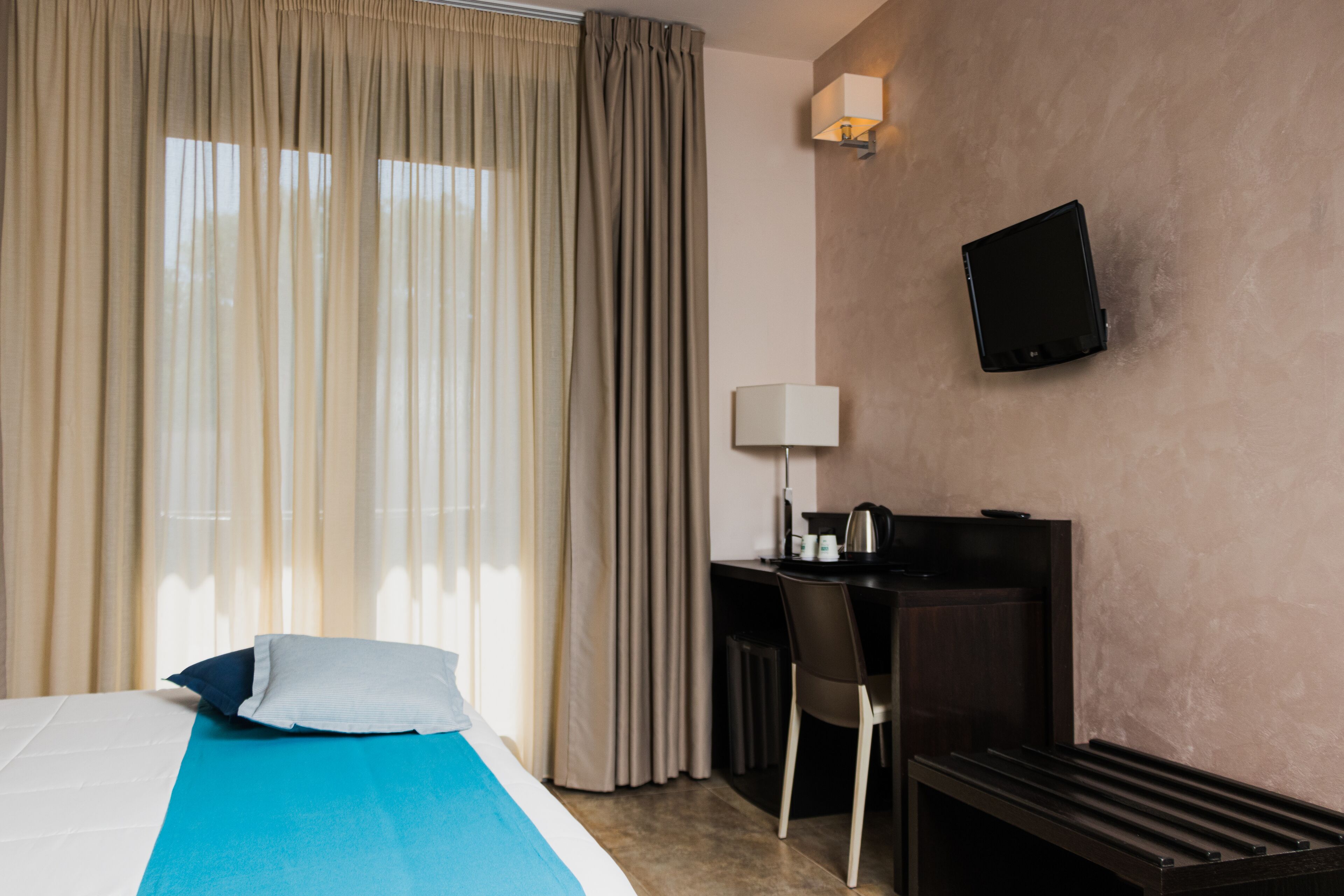 A hotel room with a bed, a desk, a chair, a TV, and a window with curtains.