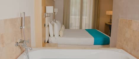 A hotel room with a large bathtub, a bed with white and teal bedding, a bedside table with a lamp, and a window with sheer curtains.