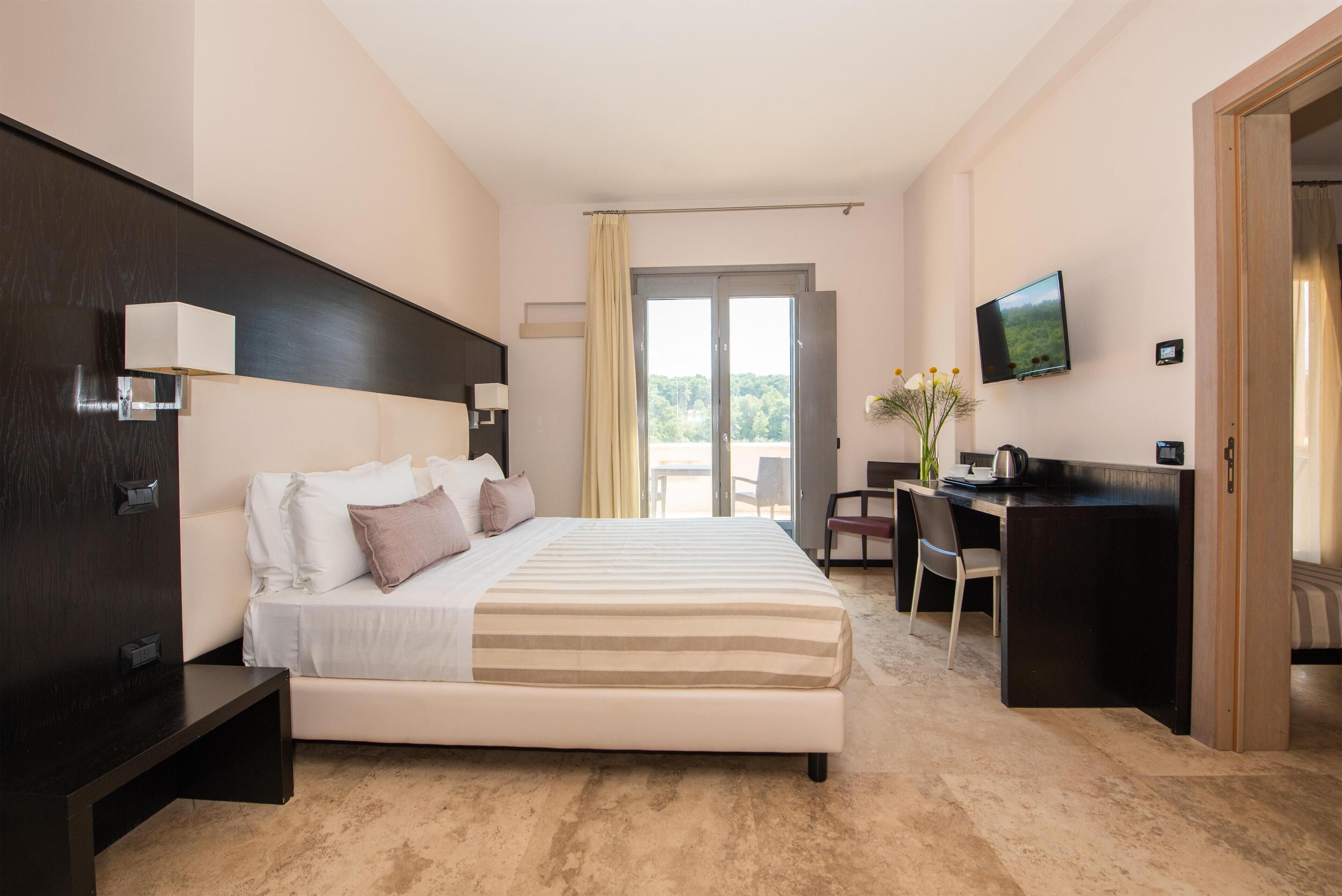 A modern hotel room with a large bed, a desk, a TV, and a balcony with a view.
