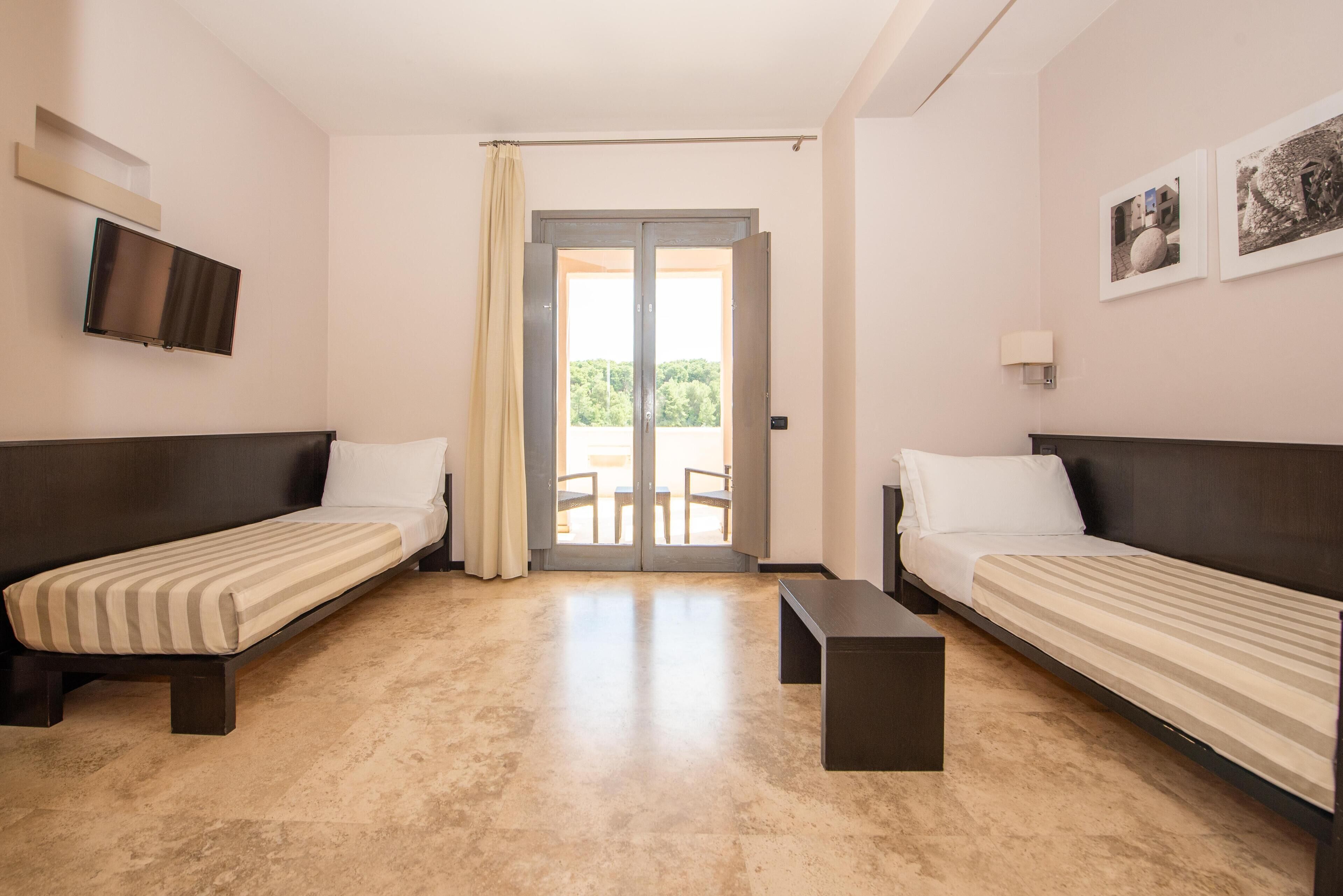 A room with two single beds, a flat-screen TV mounted on the wall, a small table, and a sliding glass door leading to an outdoor area.