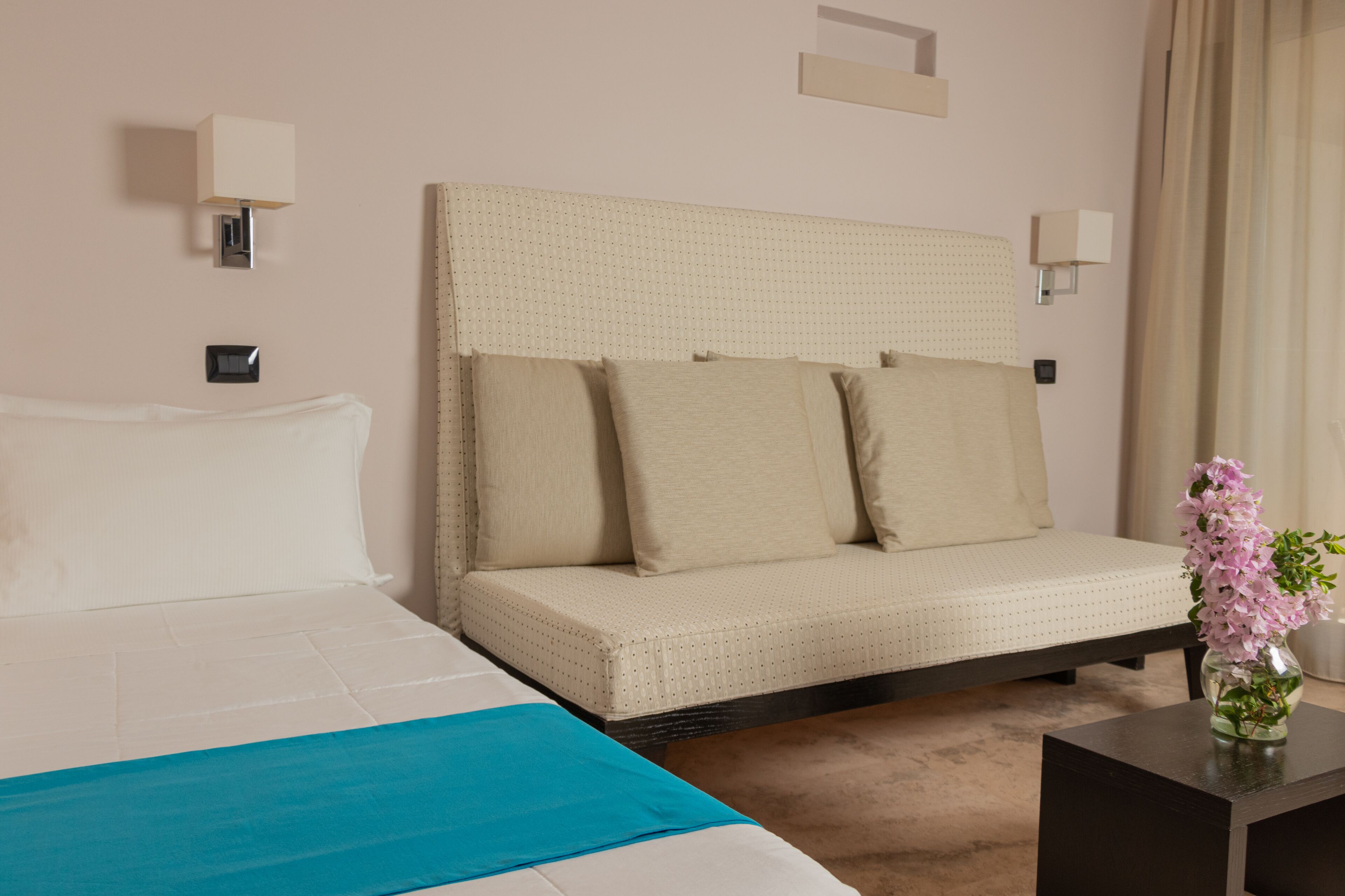 A hotel room with a large bed, a sofa bed, a small table with a vase of flowers, and two wall-mounted lamps.