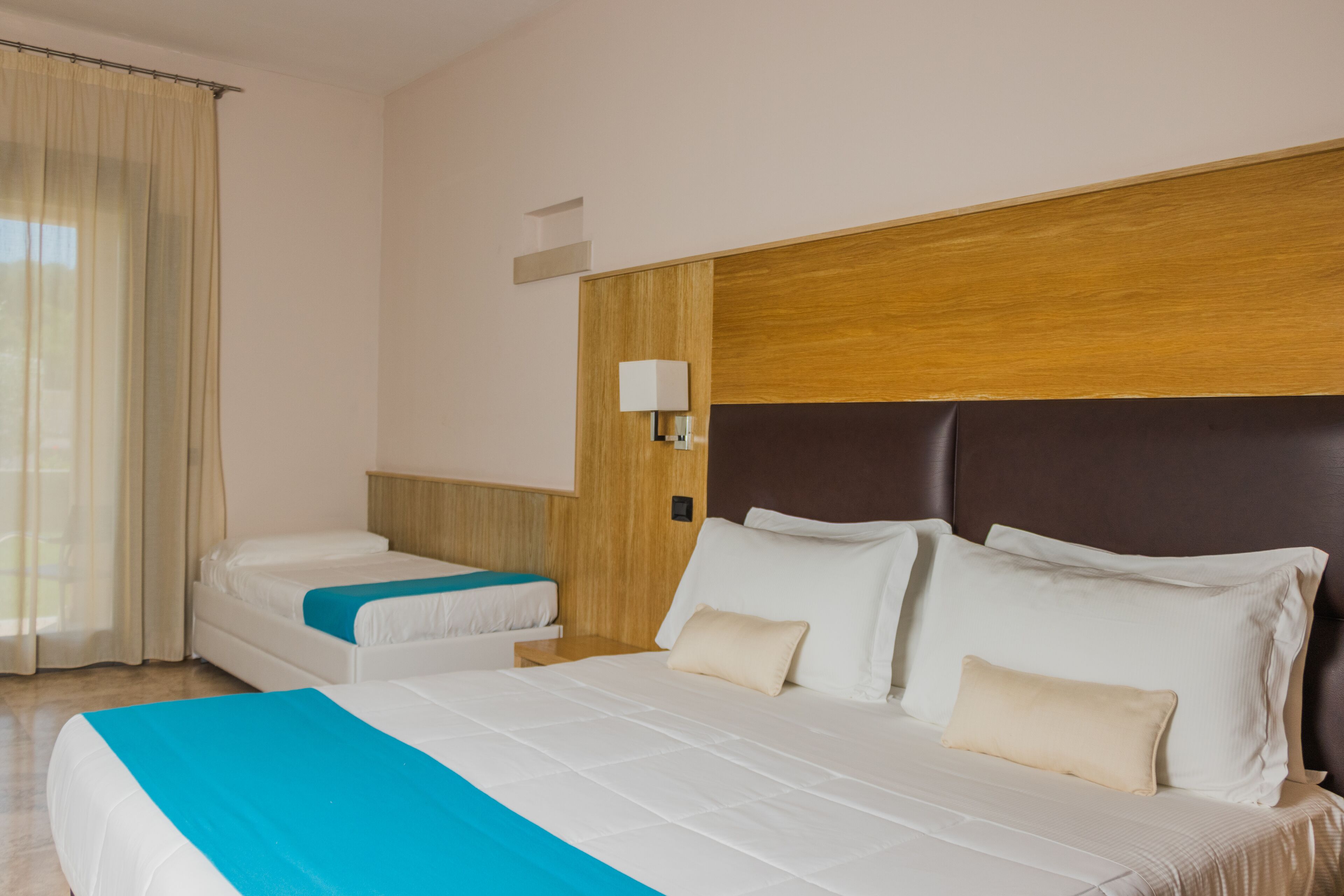 Habitación de hotel con una cama grande, una cama más pequeña, una mesita de noche, una lámpara y una ventana con cortinas.