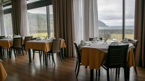 A restaurant with tables set for dining, large windows with curtains, and a view of the sea and hills. - Inatel Flores Hotel (Santa Cruz das Flores)