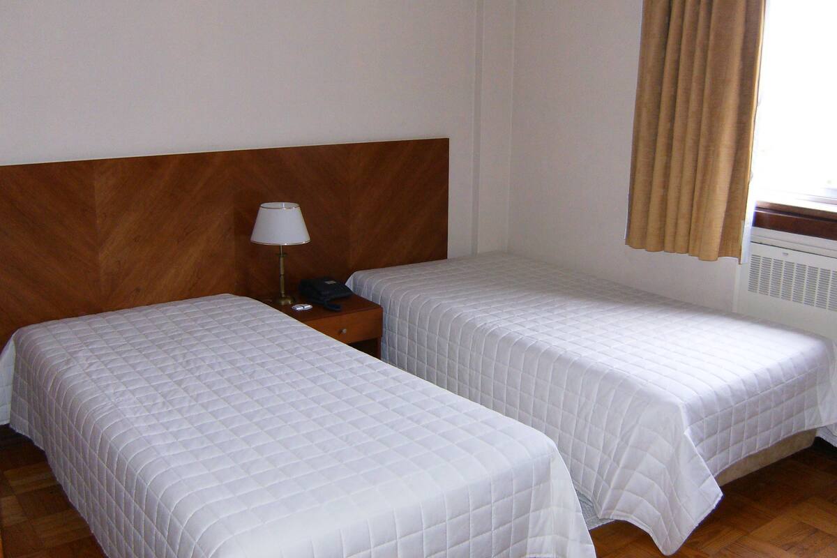 A hotel room with two single beds, a wooden headboard, a bedside table with a lamp, and a window with curtains.