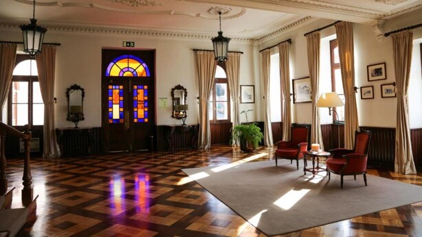 A hotel lobby with a checkered floor, a stained glass door, and a seating area with chairs and a table. - INATEL Luso (Mealhada)
