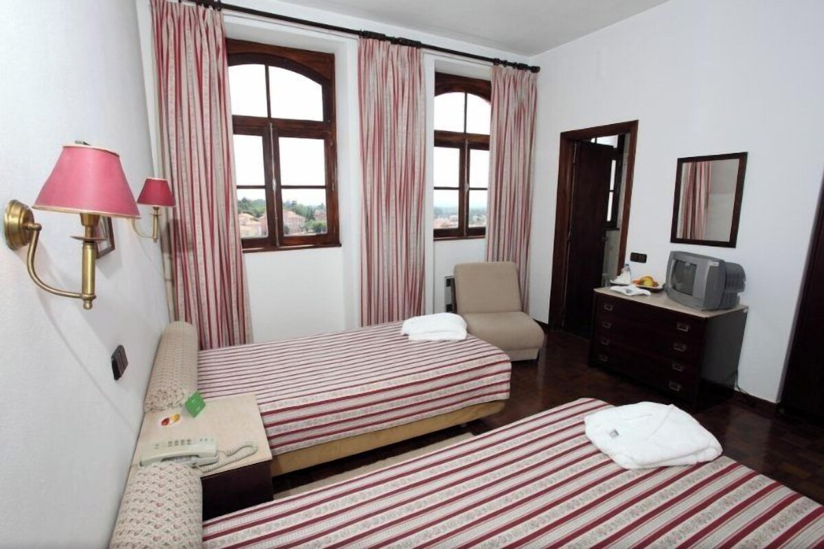 Habitación de hotel con dos camas, una silla, un tocador, un televisor y una ventana con cortinas.