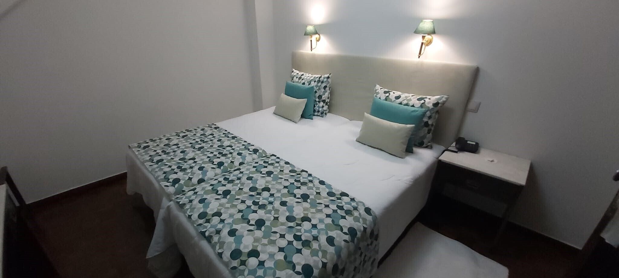 Una cama bien hecha con una colcha estampada y almohadas, dos lámparas de noche y una mesita auxiliar con toma de corriente.