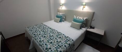 Una cama bien hecha con una colcha estampada y almohadas, dos lámparas de noche y una mesita auxiliar con toma de corriente.