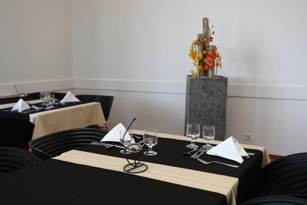 A dining area with tables set for four, each with a black tablecloth, white napkins, and clear glassware. A tall, narrow vase with orange flowers is placed on a pedestal between the tables. - INATEL Luso (Mealhada)