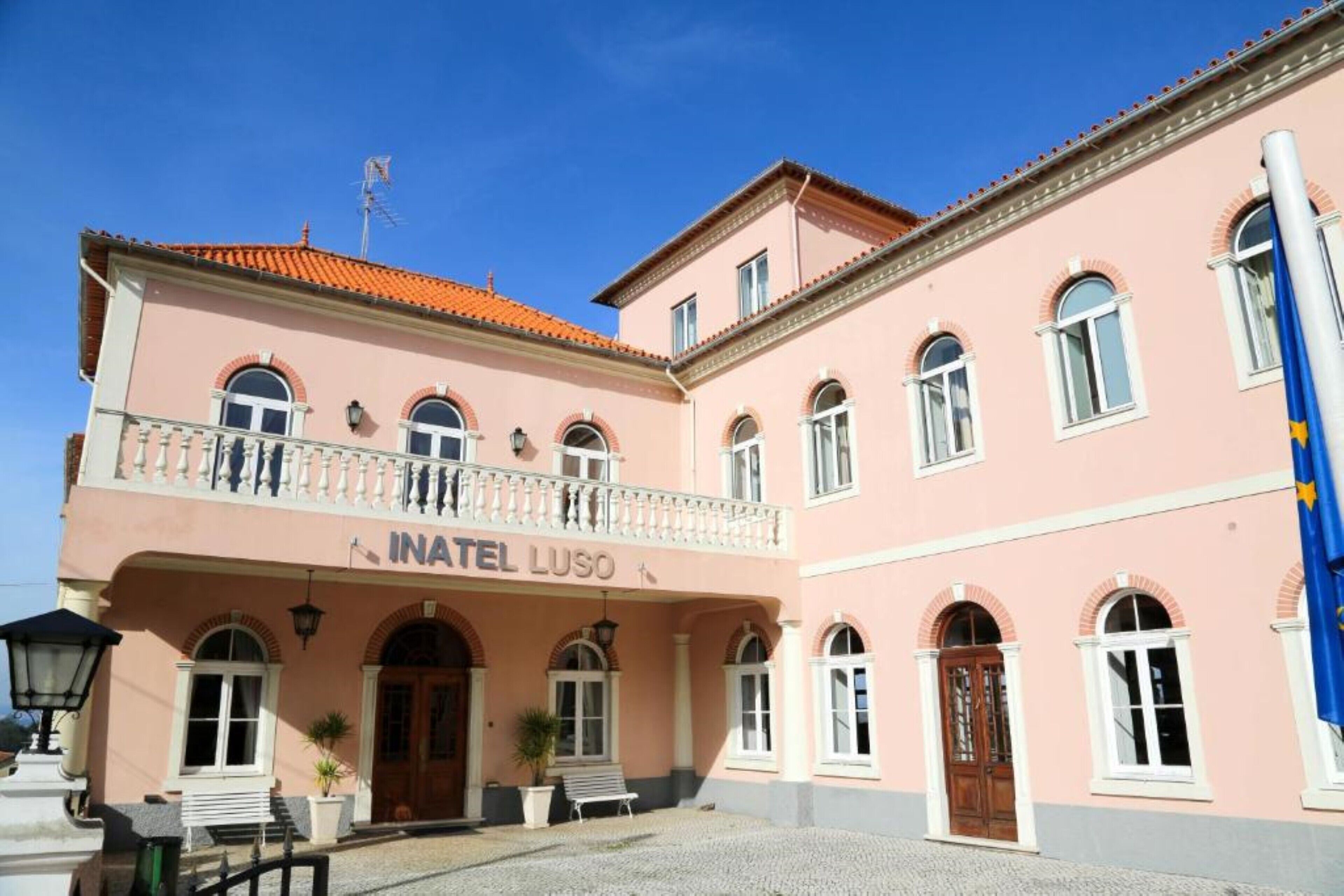 Un edificio rosa con el letrero "INATEL LUSO" en la fachada, que presenta ventanas de arco y un balcón.