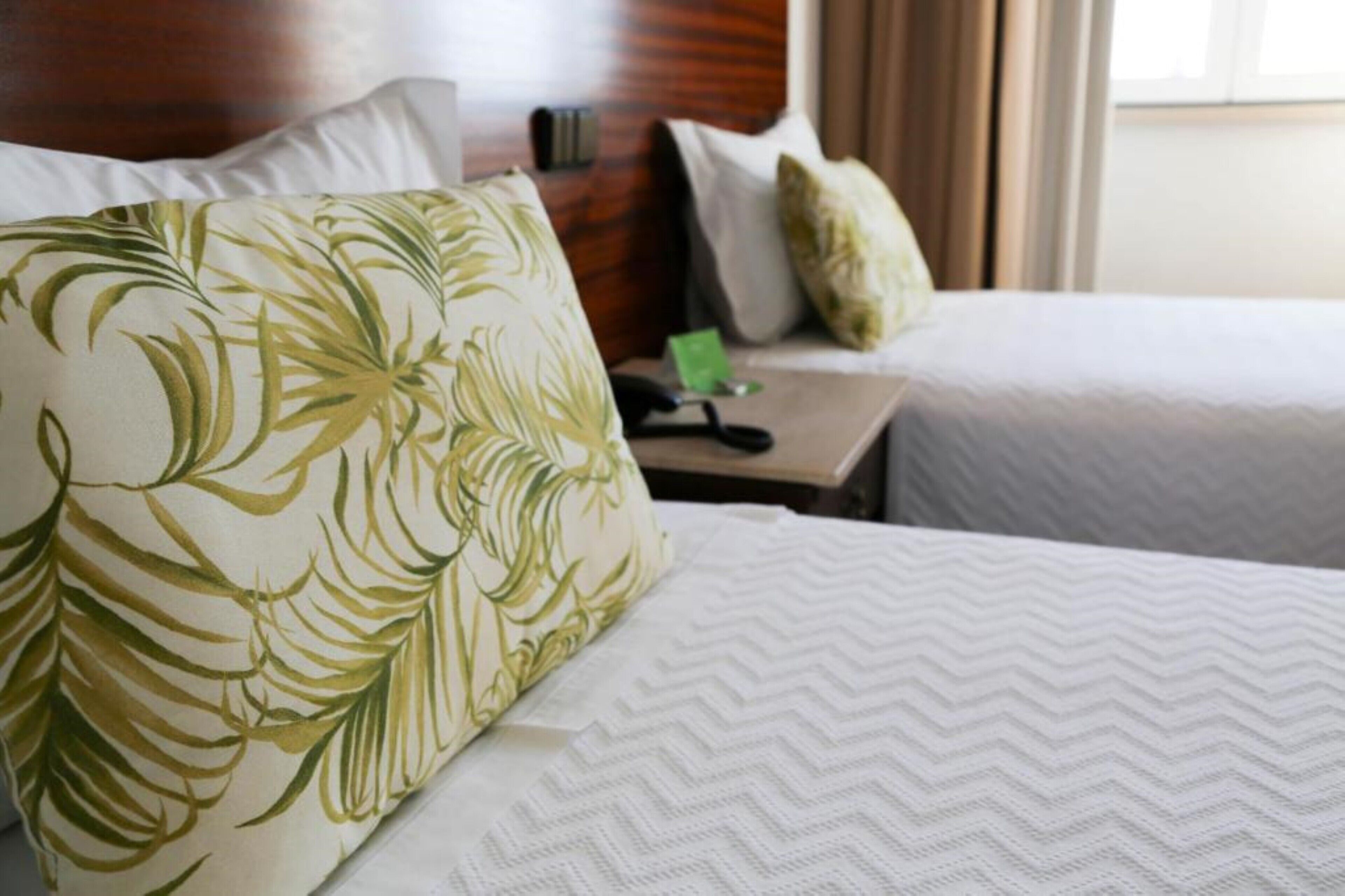 Habitación de hotel con dos camas, una con almohada estampada con motivos de hojas y la otra con una almohada lisa. Hay una mesita de noche con un teléfono y una tarjeta verde.