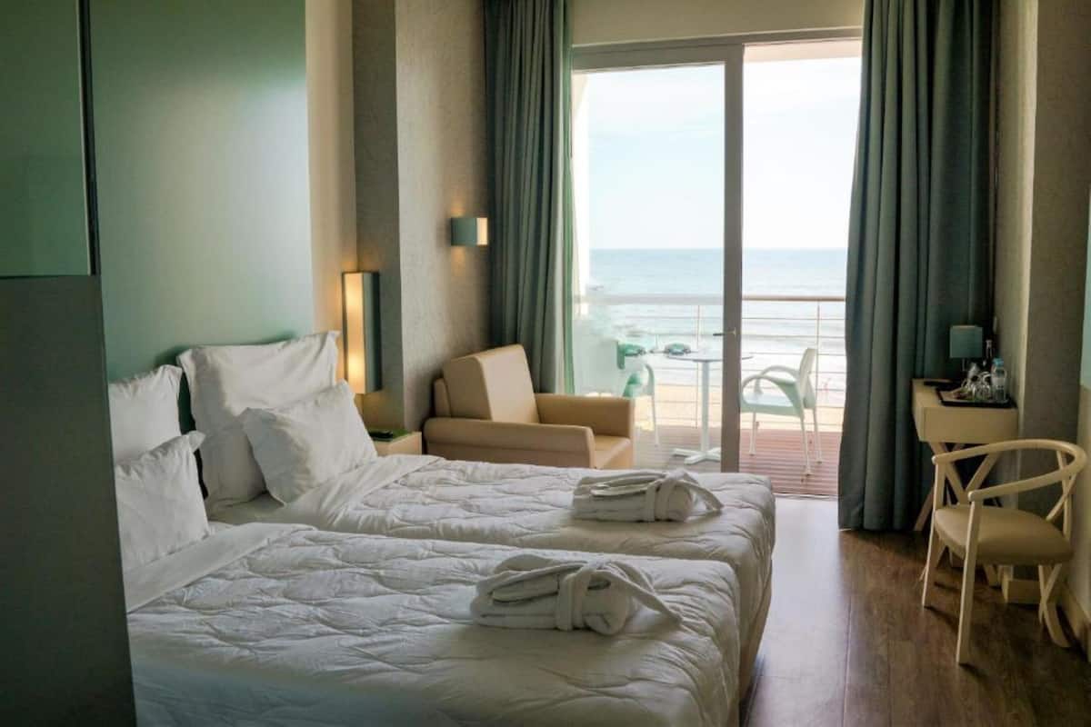 A hotel room with two beds, a sofa, a chair, and a balcony with a view of the sea.