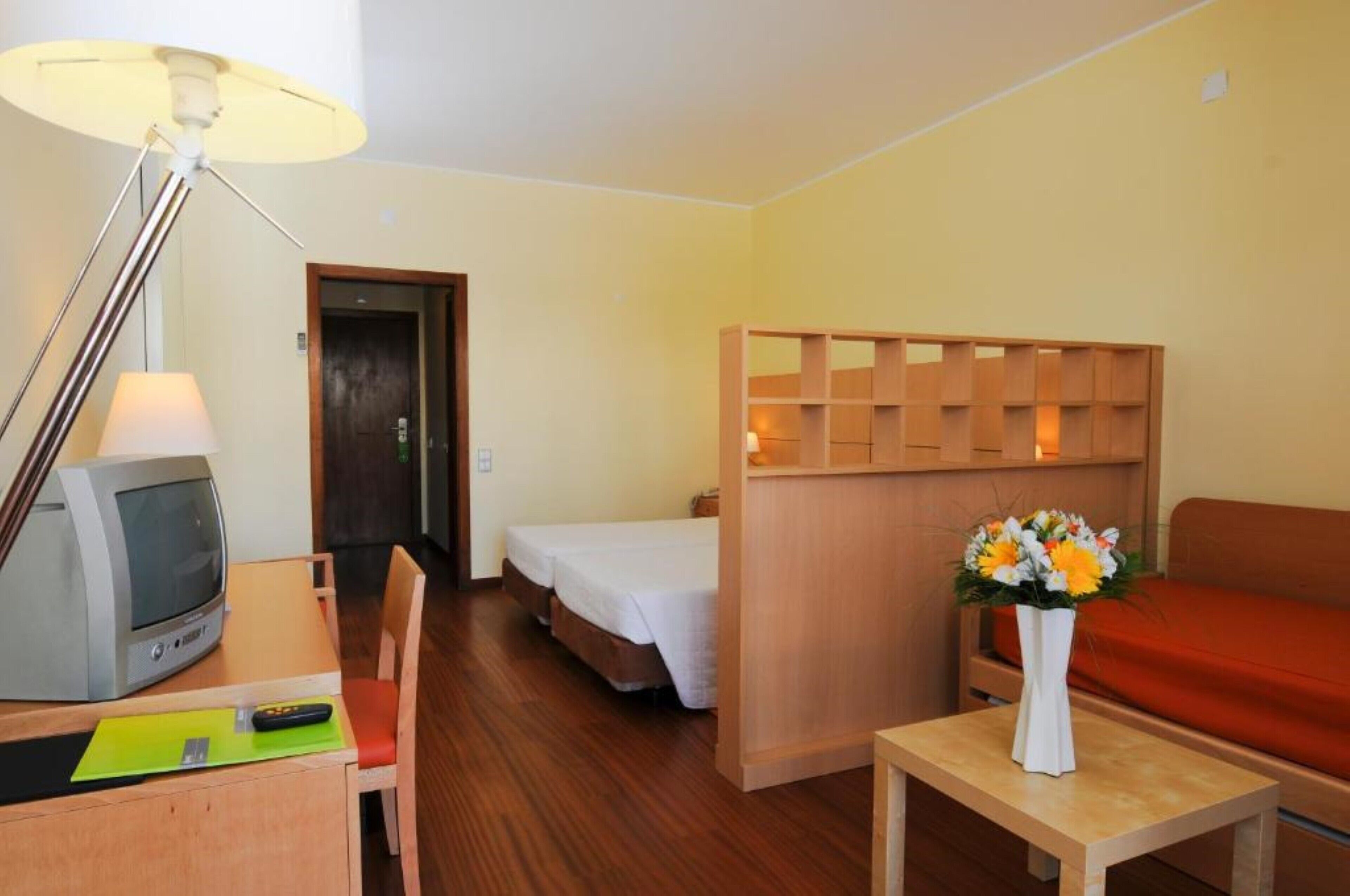 A hotel room with a wooden desk, a television, a bed, a sofa, and a small table with a vase of flowers.