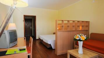 A hotel room with a wooden desk, a television, a bed, a sofa, and a small table with a vase of flowers.