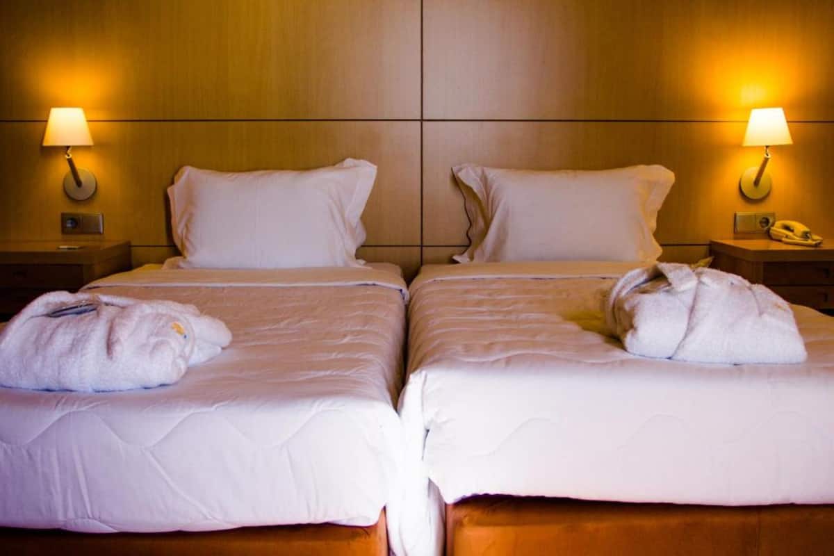 A hotel room with two beds, each with a white bedspread and pillows, and two bedside lamps. There are towels on each bed.