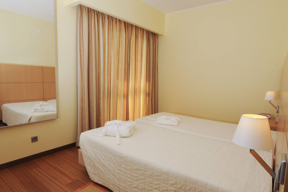 A hotel room with a bed, a mirror, a window with curtains, and a bedside lamp.
