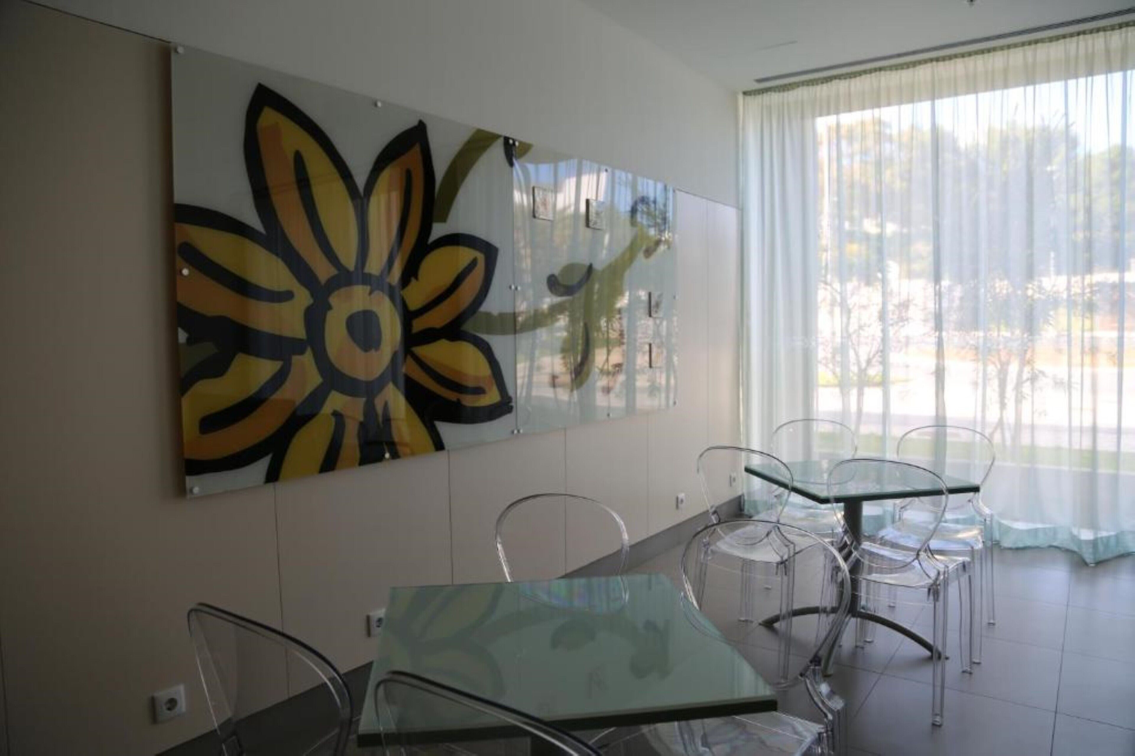 A modern dining area with glass tables and chairs, a large abstract painting on the wall, and a view through sheer curtains.